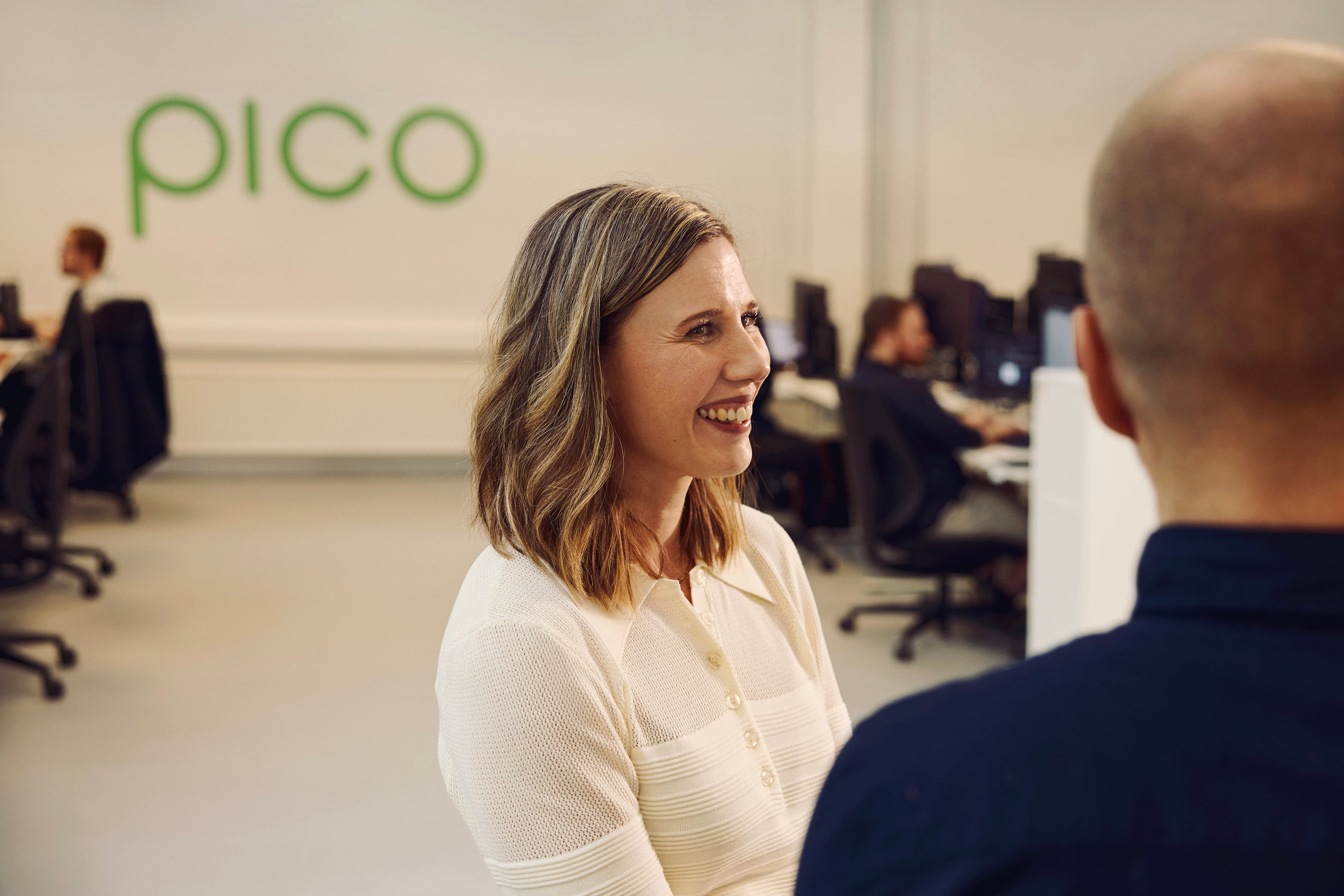 Kvinde i hvid skjorte taler med kollega på Pico-kontor med medarbejdere i baggrunden