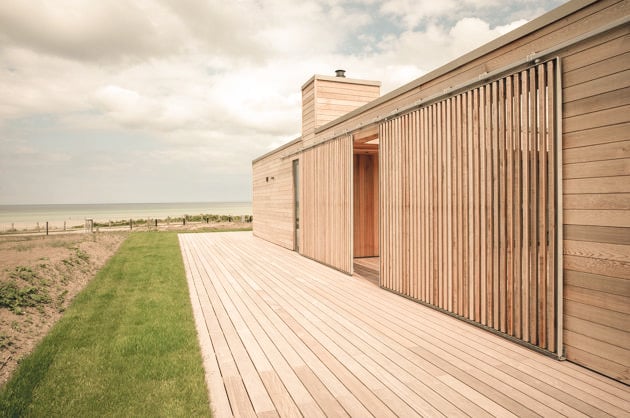 Moderne træbygning med terrasse ved strand, udsigt over marker og hav