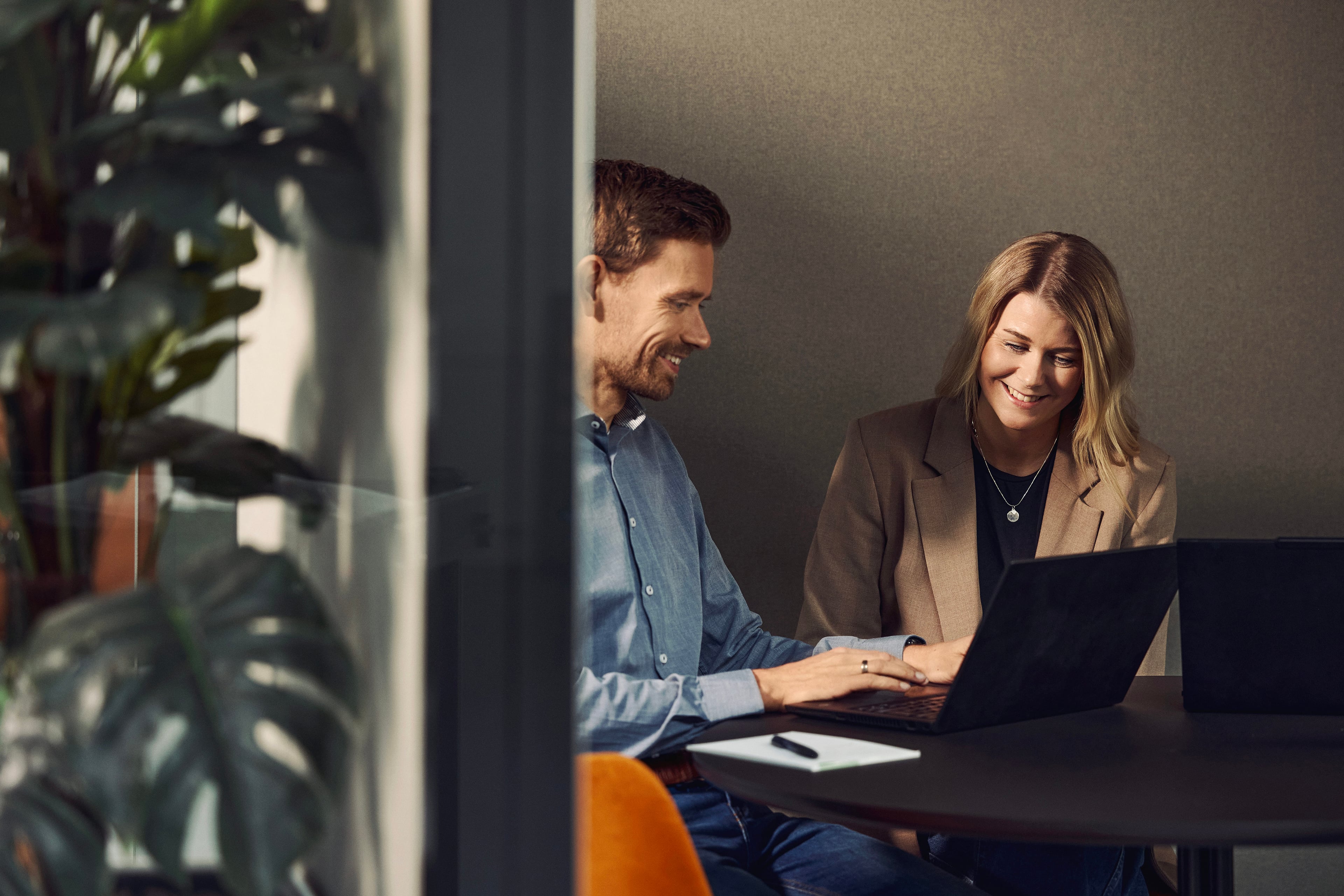 To personer arbejder sammen ved en laptop ved et bord på et kontor med planter i baggrunden.