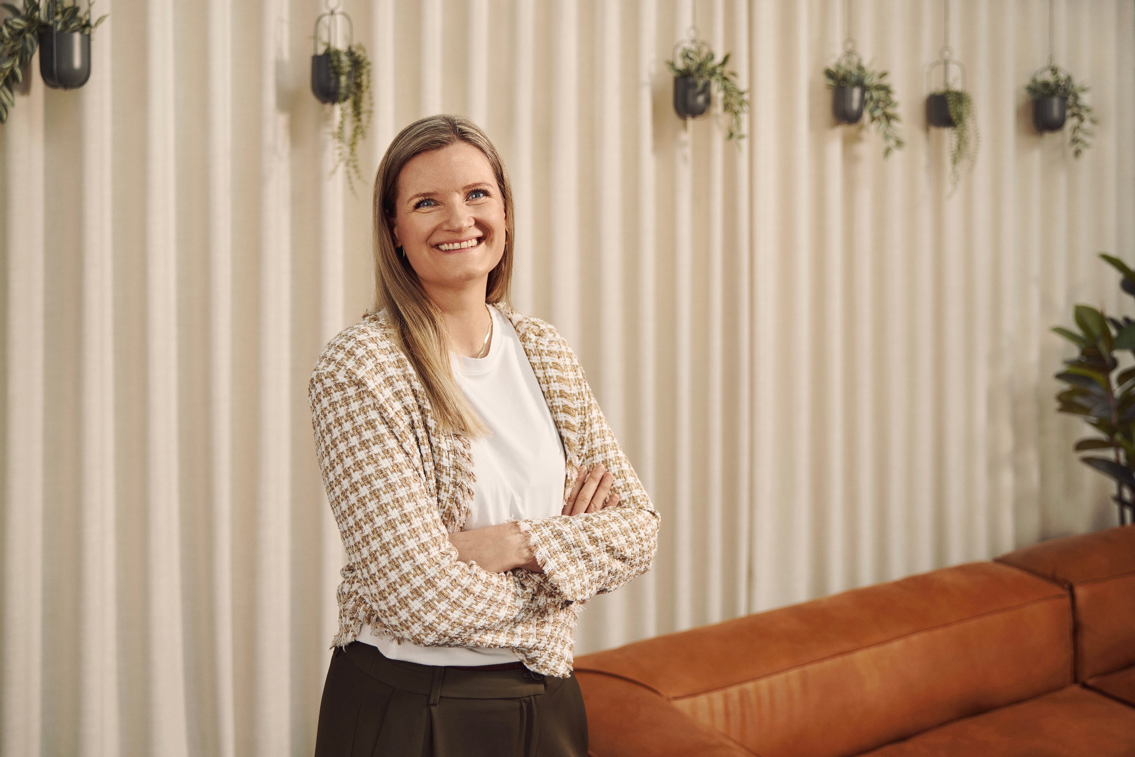 Kvinde i beige blazer står foran stribet væg med hængende planter og lædersofa