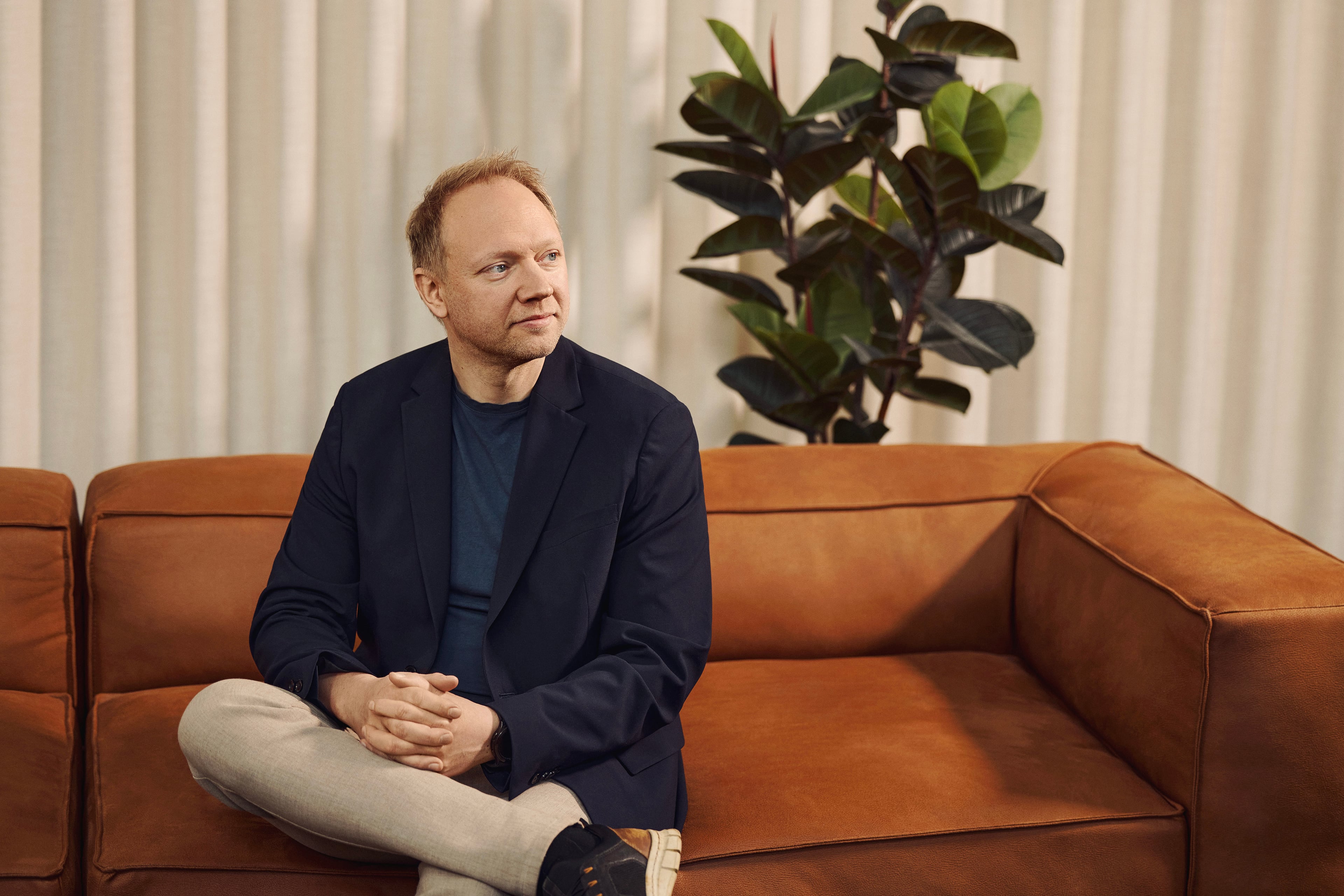 Mand i marineblå blazer sidder på cognacfarvet sofa foran hvide gardiner og grøn plante