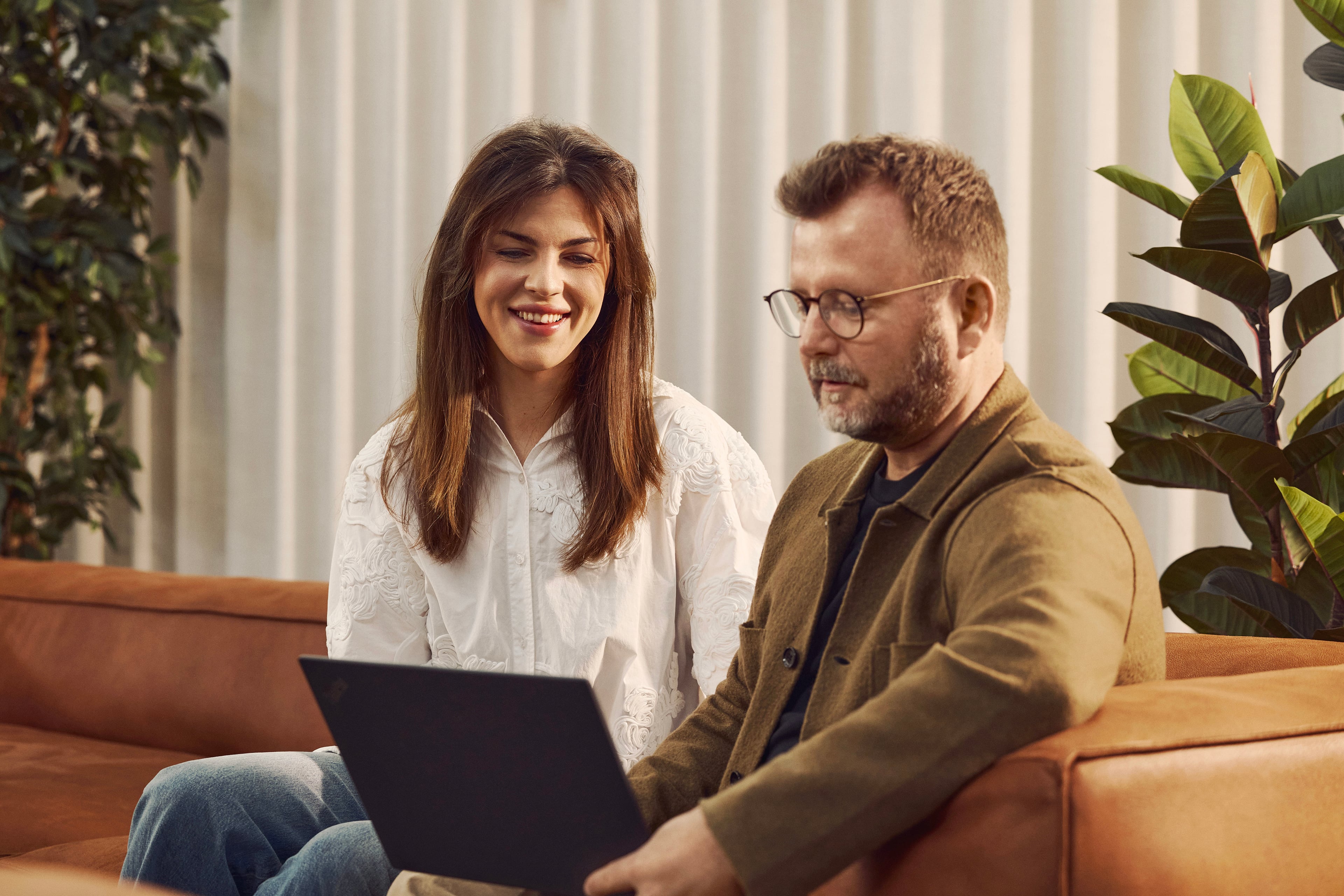 To personer sidder på sofa med bærbar computer. Kvinde i hvid skjorte og mand i brun jakke arbejder sammen ved laptop.
