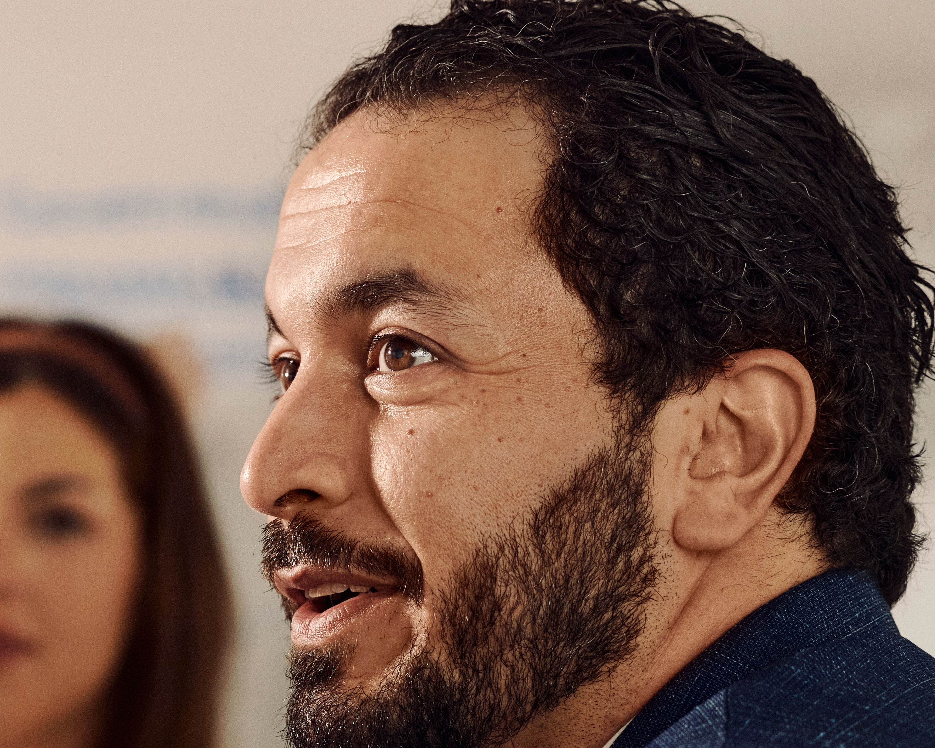Mand med skæg i profil, ser til siden med udtryksfuldt ansigt. Anden person uskarp i baggrunden.