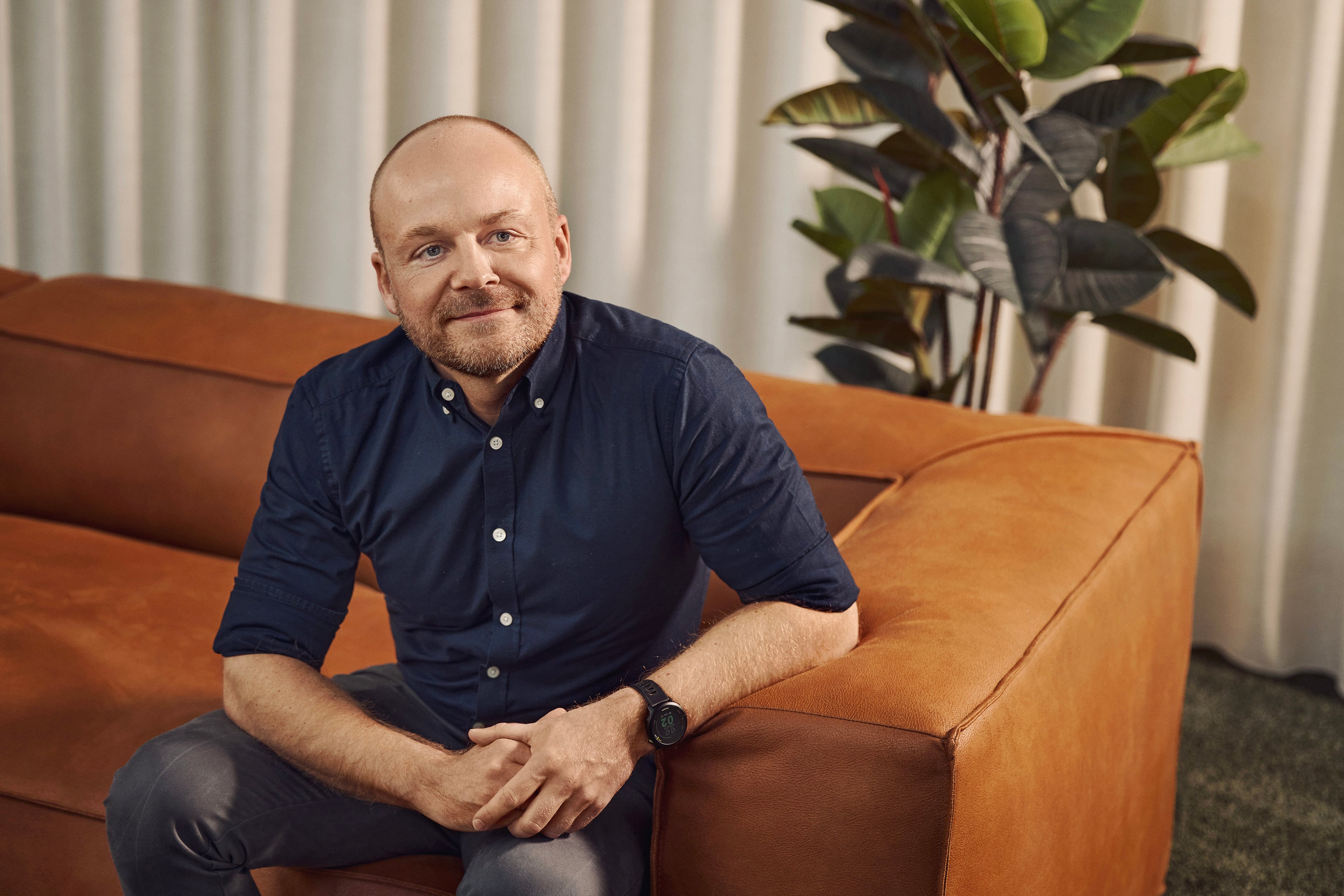 Mand i marineblå skjorte sidder på cognacfarvet sofa foran hvide gardiner og grøn plante.
