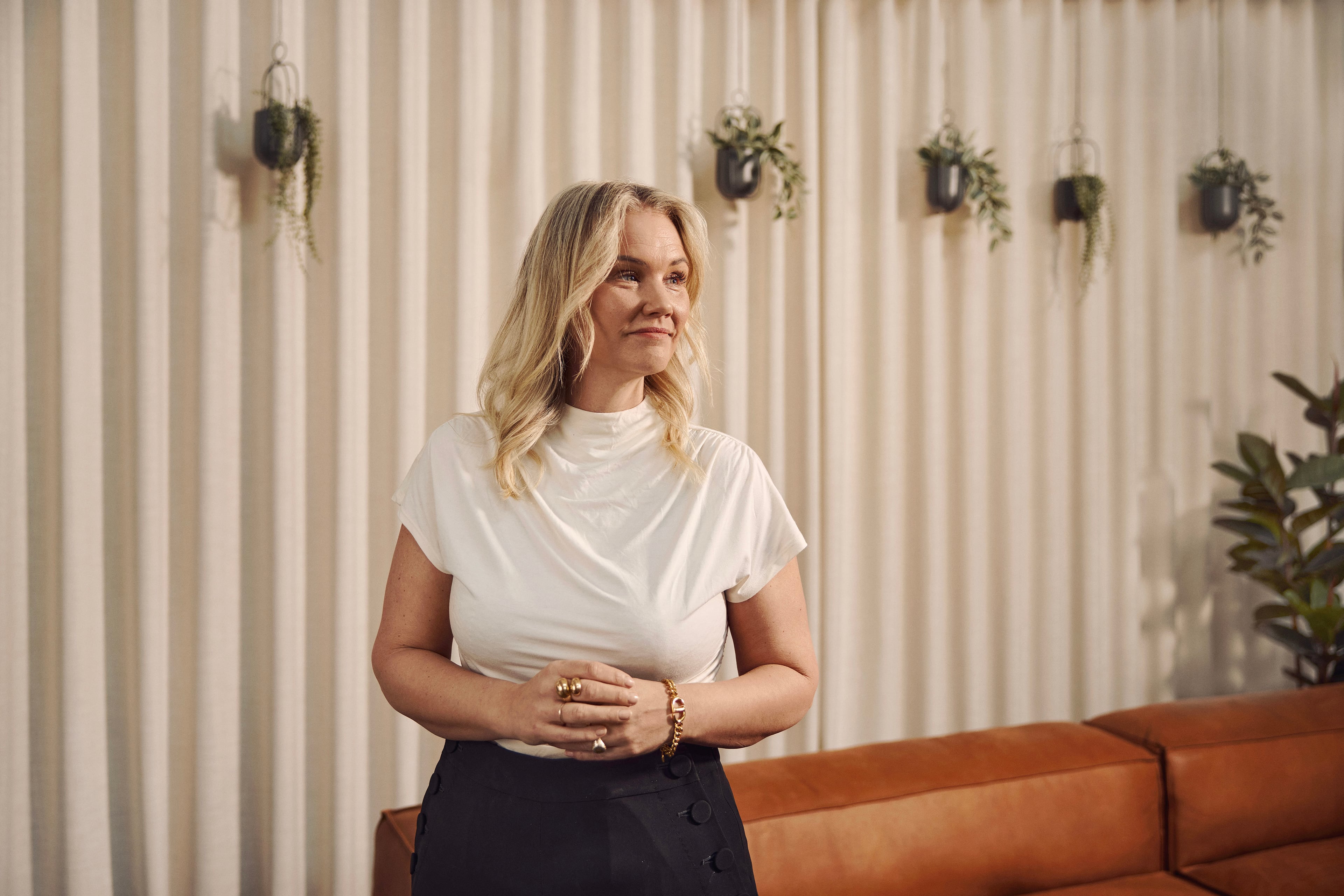 Kvinde i hvidt t-shirt og mørk nederdel står foran stribet væg med hængende planter